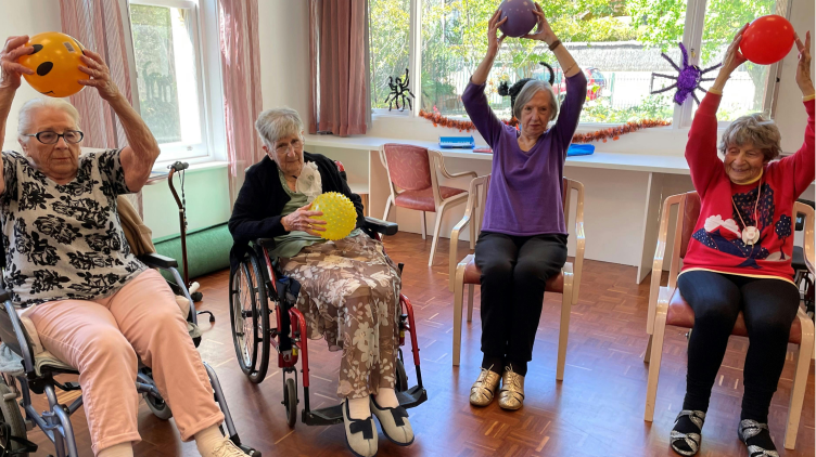 Group of ladies sitting in chairs exercising with balloons and balls holding them above their heads.