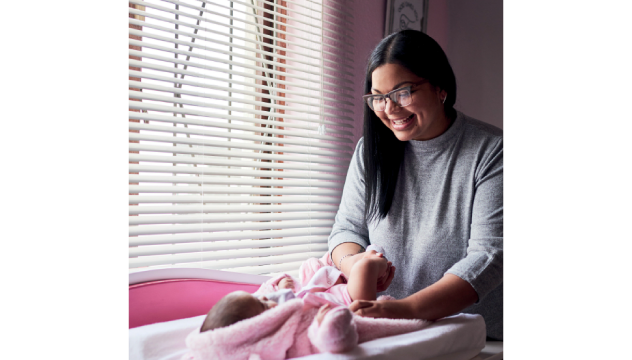 Woman with black hair, wearing a grey jumper and glasses standing with a baby lying on a changing mat